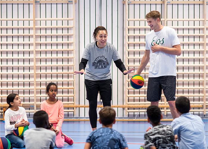Två lärare står i en ring med barn. Kvinnan gestikulerar med händerna och mannen dribblar med en basketboll bredvid henne.