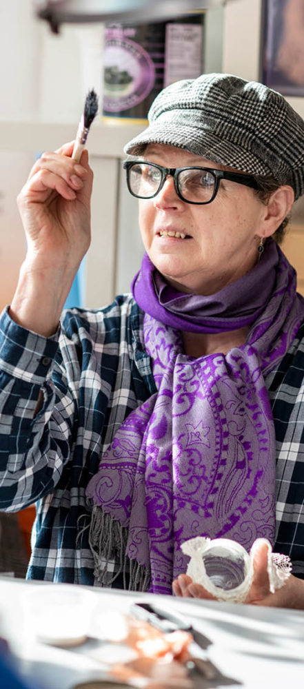 Kvinna med keps, glasögon och lila sjal runt halsen sitter med en pensel i högsta hugg.