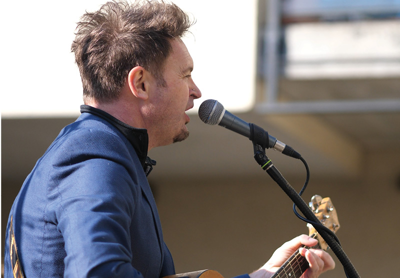 Porträtt från sidan på en man som sjunger i en mikrofon och spelar gitarr. Solen skiner.
