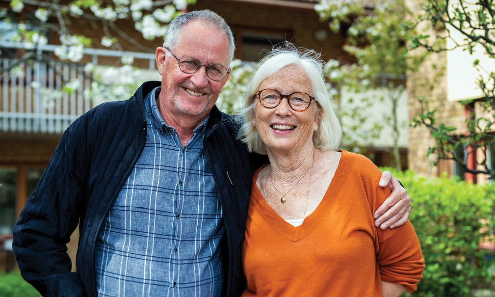 En man och kvinna i 60-årsåldern som glada tittar in i kameran och håller om varandra. I bakgrunden syns en lummig innergård.