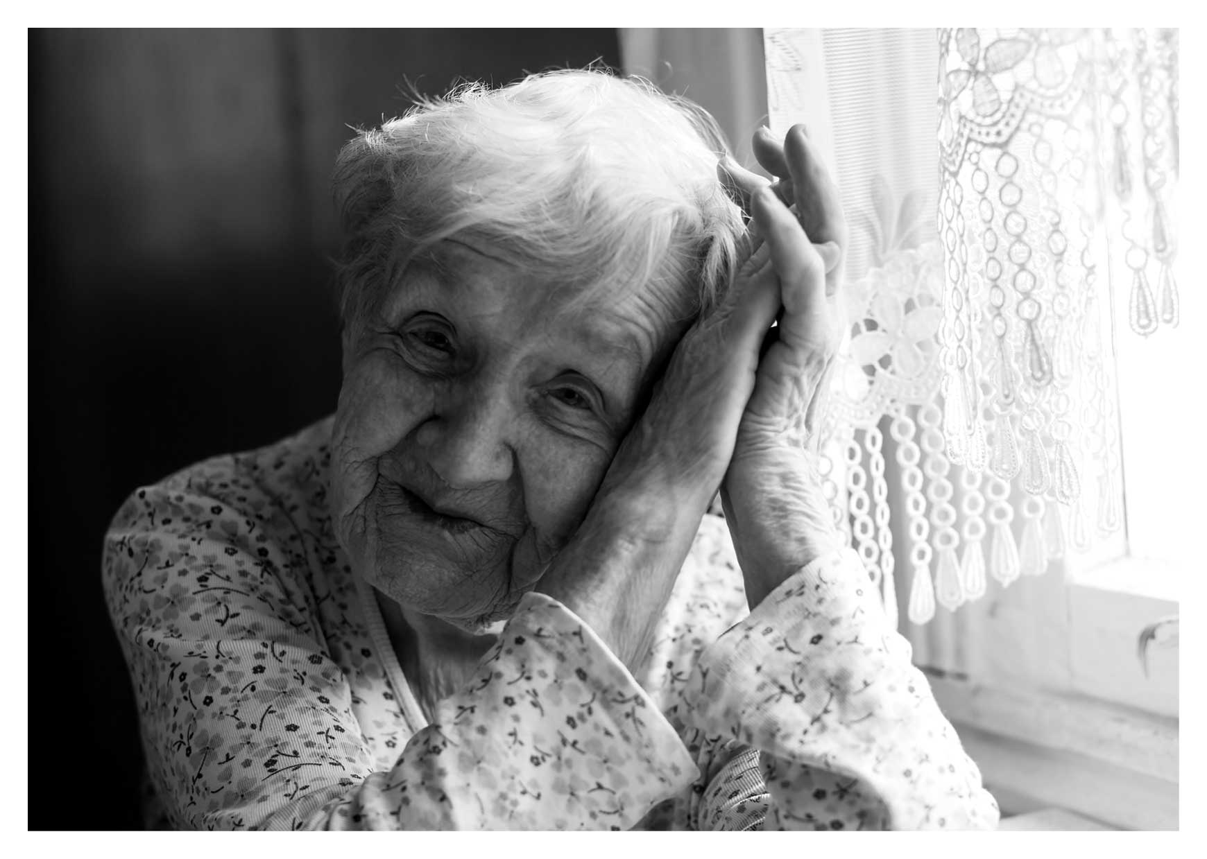 Svartvit porträttbild av en äldre kvinna i hemmamiljö, hon ser ut att sitta vid ett köksbord med gardin i bakgrunden.
